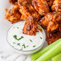 A buffalo chicken wing dipped in a bowl of blue cheese dressing, surrounded by more buffalo wings and celery sticks on a plate.