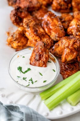 A buffalo chicken wing dipped in a bowl of blue cheese dressing, surrounded by more buffalo wings and celery sticks on a plate.