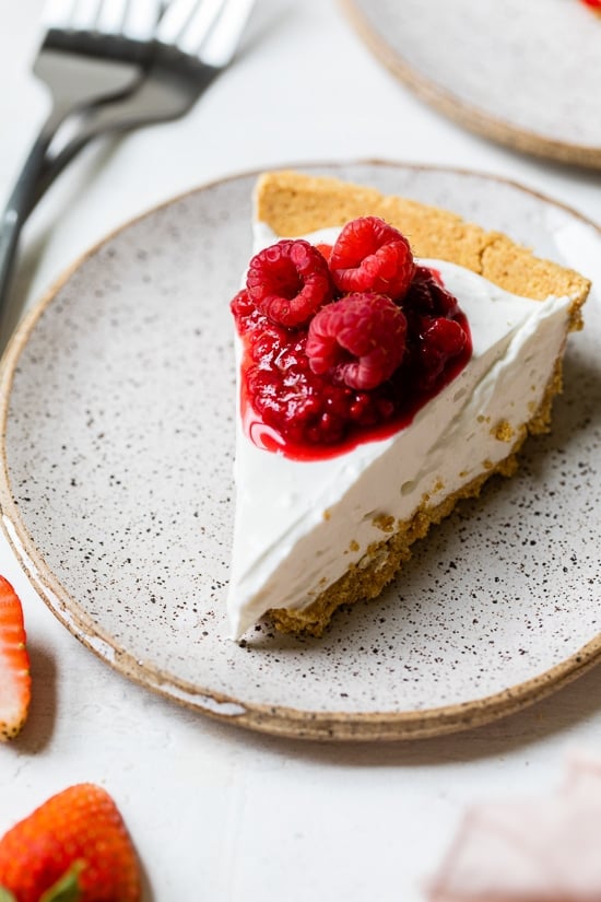 This easy no-bake cheesecake is light, dreamy and so easy to make, you don’t even need an oven. A little slice of heaven! Slice of no-bake cheesecake on plate with macerated and fresh raspberries on top