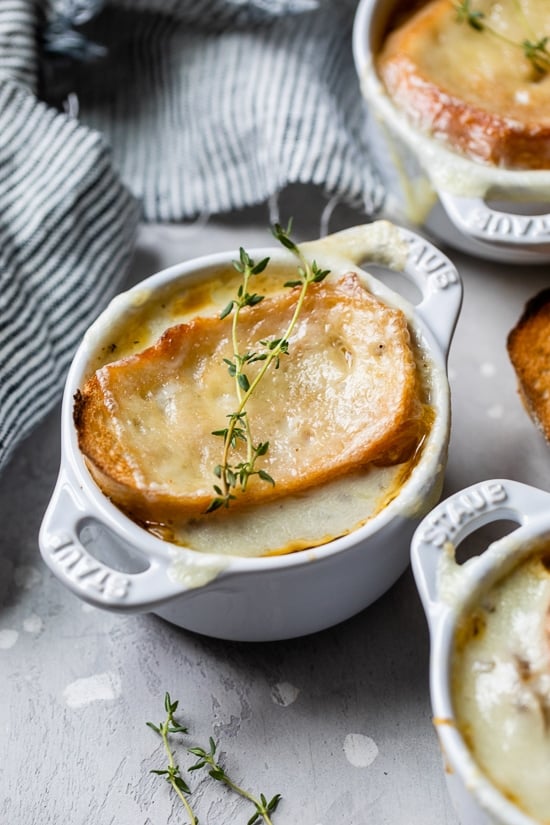 This lightened-up French onion soup recipe is just as delicious as the original, with beefy broth, caramelized onions, and melty cheese! French onion soup in ramekins topped with toast and melted cheese, garnished with thyme sprigs.