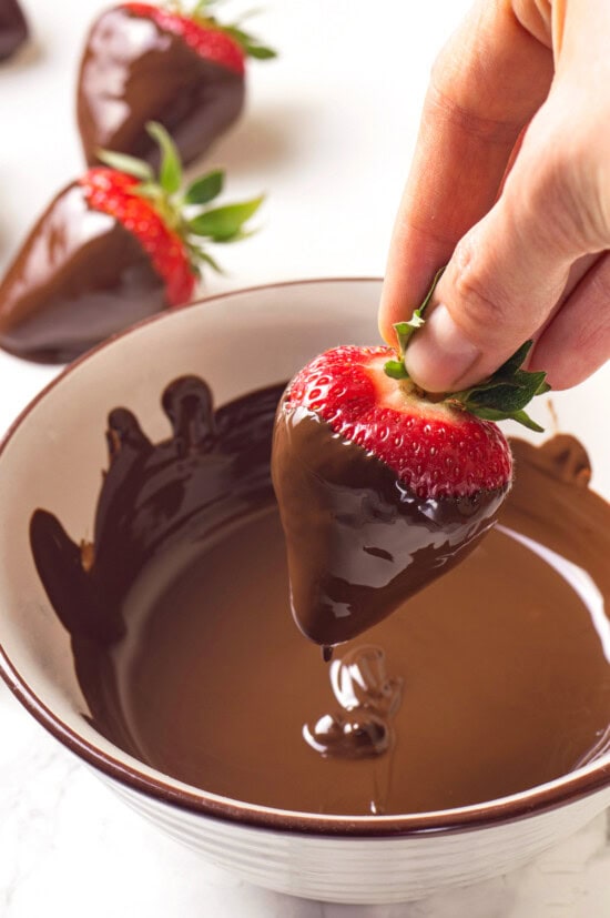 Chocolate-covered strawberries are an elegant treat that's incredibly easy to make at home. You'll never buy them at the store again! Hand dipping strawberry into bowl of melted chocolate