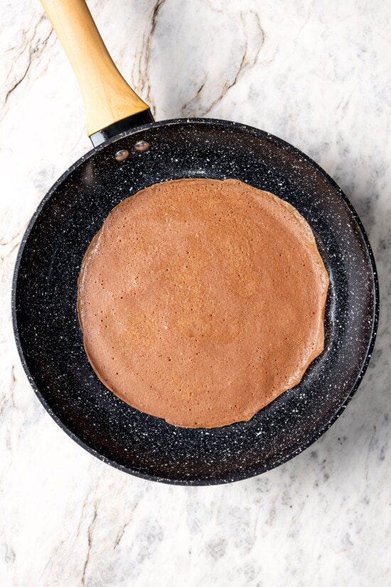 These chocolate crepes are thin, tender and lightly sweet, filled with strawberries and whipped topping. Serve them for breakfast or dessert! Chocolate crepe in skillet.