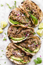 5 pork carnitas tacos topped with slices of avocado and red onion.
