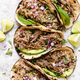 5 pork carnitas tacos topped with slices of avocado and red onion.