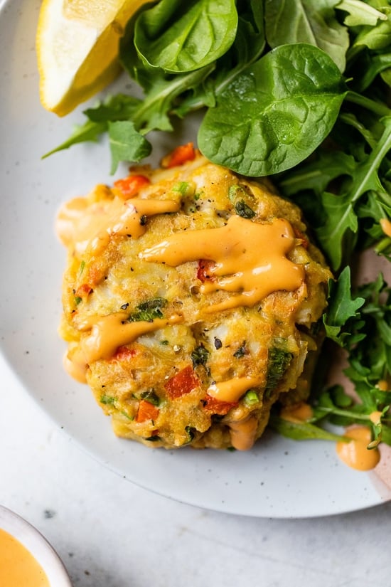 This easy baked crab cakes recipe combines juicy lump crab meat, simple seasonings, and fresh herbs. Perfect as an appetizer or light dinner! A baked lump crab cake drizzled with red pepper sauce next to a green salad on a plate.