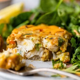 One half of a baked crab cake next to a fork on a plate, with a green salad in the background.