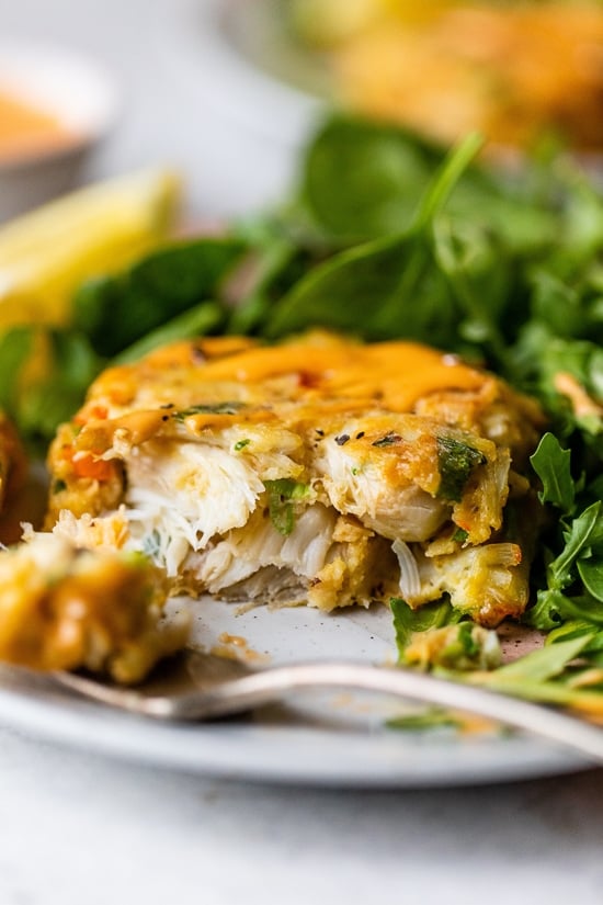 This easy baked crab cakes recipe combines juicy lump crab meat, simple seasonings, and fresh herbs. Perfect as an appetizer or light dinner! One half of a baked crab cake next to a fork on a plate, with a green salad in the background.