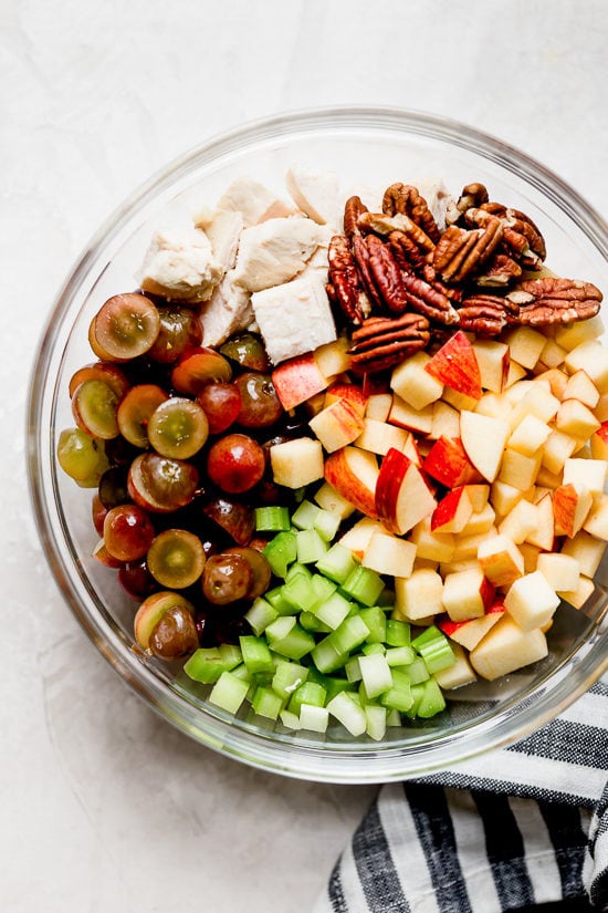 This lightened up chicken Waldorf salad is creamy and crisp, with tender chicken, apples, grapes, celery and nuts in a light dressing. Ingredients for chicken Waldorf salad in glass bowl.