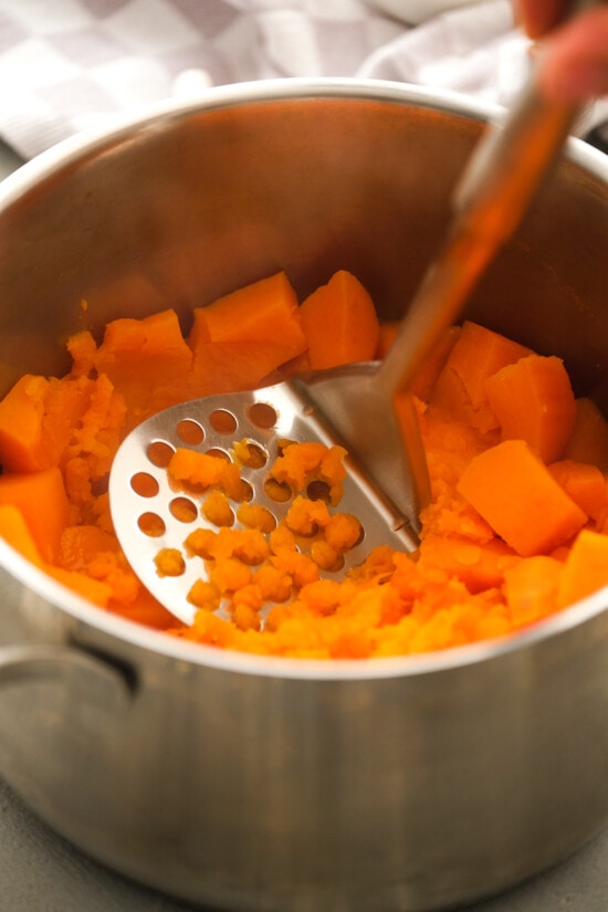 This lighter sweet potato casserole is made with golden raisins, crushed pineapple, and spices, topped with toasty marshmallows and pecans. A potato masher being used to mash cooked sweet potatoes in a pot.