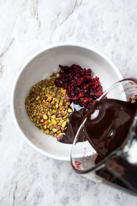 This easy Dark Chocolate Bark with Pistachios and Cranberries is festive enough for a holiday treat, but healthy enough to enjoy year round! Pouring melted chocolate into bowl with pistachios and cranberries.