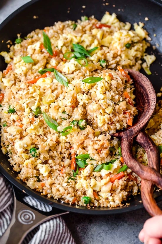 This low-carb cauliflower fried rice recipe is quick, flavorful and a great way to enjoy your favorite takeout-style dish in a lighter way! Skillet of cauliflower fried rice with wooden spoon.