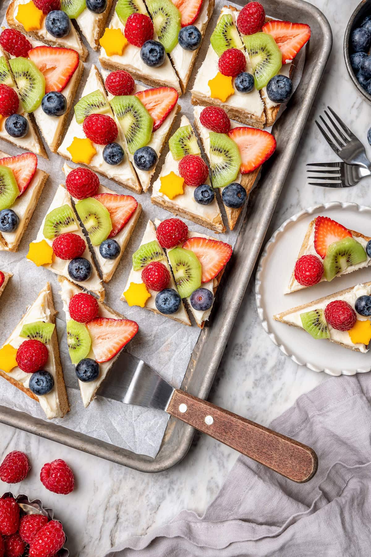 This easy fruit pizza is made with a lightened-up sugar cookie crust and topped with fluffy cream cheese frosting and colorful fresh fruit! Overhead view of fruit pizza on sheet pan and plate