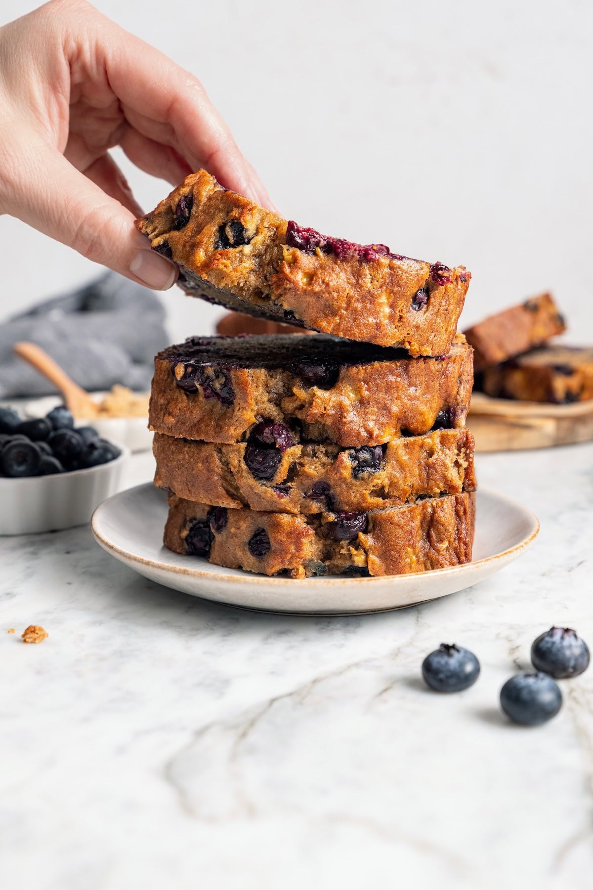 This blueberry banana bread is a lighter twist on a classic that’s perfect for breakfast, snacking, or anytime you have ripe bananas to use! Lifting slice of blueberry banana bread from plate with stack of slices.