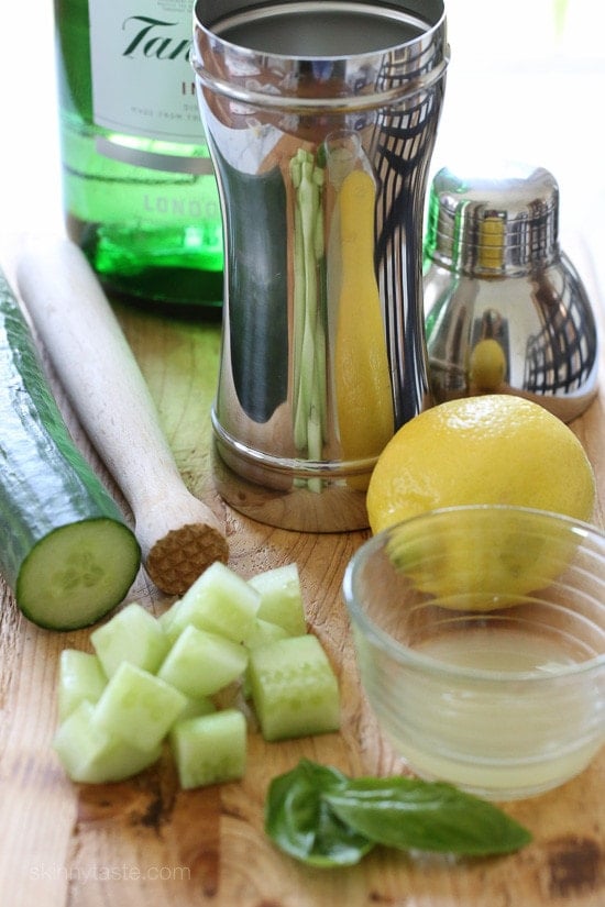Basil-Cucumber Gin Cooler A cocktail shaker, a muddler, a whole cucumber with diced pieces, a whole lemon and a bowl of lemon juice, basil leaves, and a bottle of gin in the background