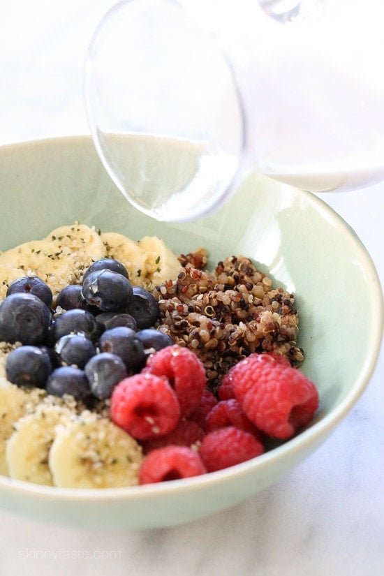 A bowl with quinoa, sliced bananas, fresh raspberries, fresh blueberries topped with hemp seeds and a pitcher pouring creamy honey drizzle on top