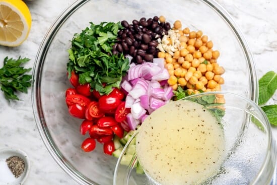 This easy garbanzo bean Mediterranean salad is fresh, simple, and full of flavor, made with minimal prep and wholesome ingredients. Pouring lemon dressing into bowl with bean salad ingredients.