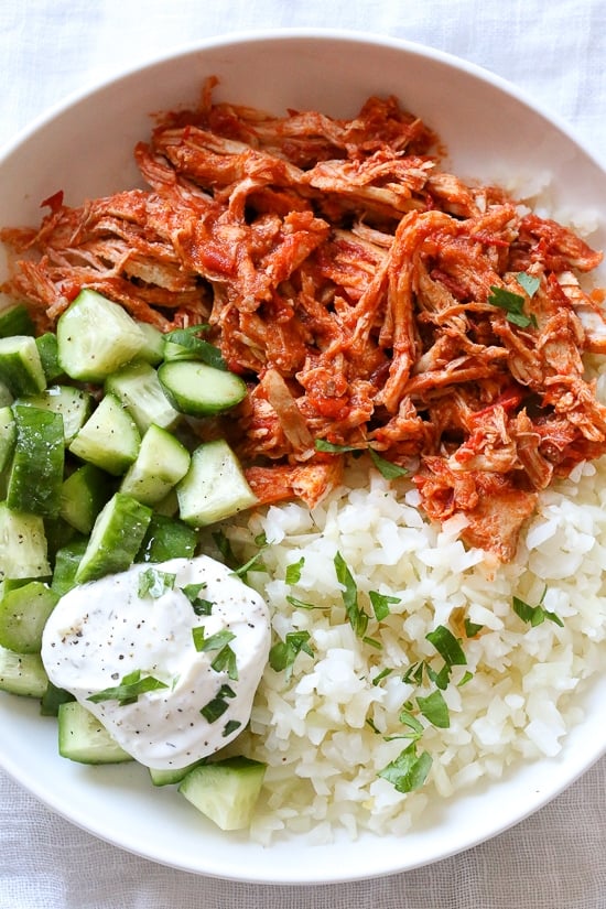 An easy harissa chicken recipe made in the slow cooker or Instant Pot, made with just 5 ingredients! Perfect for bowls, meal prep, and more. Overhead view of a harissa chicken bowl with yogurt sauce, cucumber, and rice.