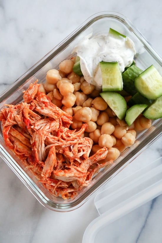 An easy harissa chicken recipe made in the slow cooker or Instant Pot, made with just 5 ingredients! Perfect for bowls, meal prep, and more. Overhead view of harissa chicken with yogurt sauce, cucumber, and rice in a meal prep container.