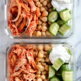 Overhead view of harissa chicken with yogurt sauce, cucumber, and rice in two meal prep containers.