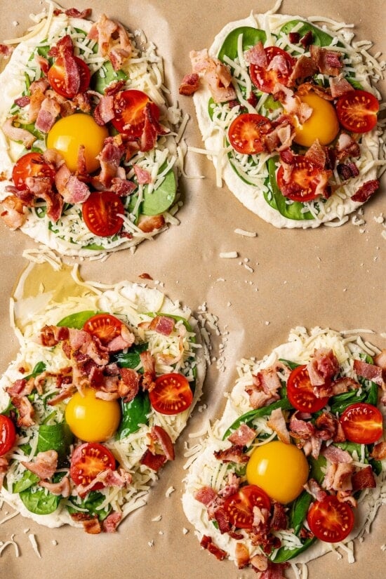 This easy breakfast pizza features a crispy yogurt crust topped with eggs, cheese, spinach and bacon for a savory protein-packed meal. Unbaked breakfast pizzas on parchment paper.