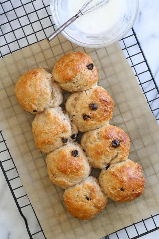 Make soft homemade hot cross buns without yeast, filled with spice, raisins, and finished with icing. Easy to bake in the oven or air fryer! Overhead view of baked hot cross buns on a parchment-lined rack next to a bowl of glaze with a whisk.