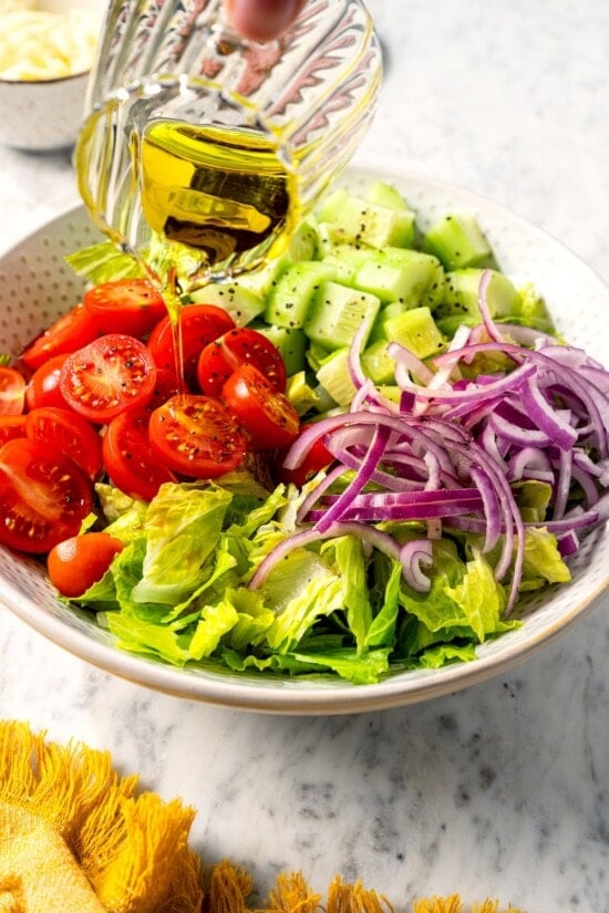 This easy Italian chopped salad pairs crisp romaine with salami, mozzarella, roasted red pepper, and veggies in red wine vinaigrette. Delish! Pouring olive oil over vegetables for Italian chopped salad.
