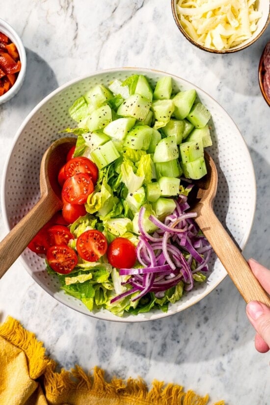 This easy Italian chopped salad pairs crisp romaine with salami, mozzarella, roasted red pepper, and veggies in red wine vinaigrette. Delish! Two spoons tossing Italian chopped salad in bowl.