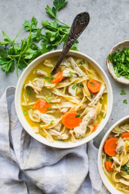 Overhead view of a bowl of chicken noodle soup next to fresh parsley and a second bowl of soup.