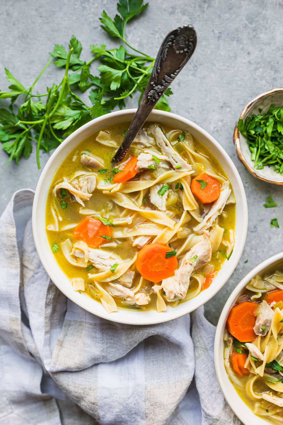 Overhead view of a bowl of chicken noodle soup next to fresh parsley and a second bowl of soup.