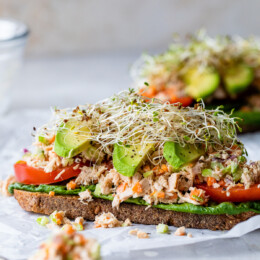 An open-faced tuna salad sandwich with avocado, with a forkful of canned tuna in the foreground.