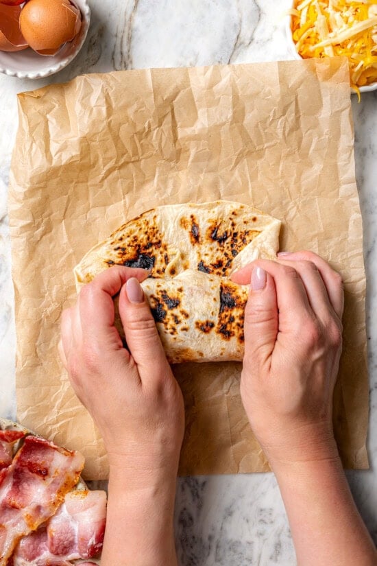 These easy freezer breakfast burritos are a delicious, protein-packed way to start the day with scrambled eggs, veggies, bacon and cheese. Folding breakfast burrito in tortilla