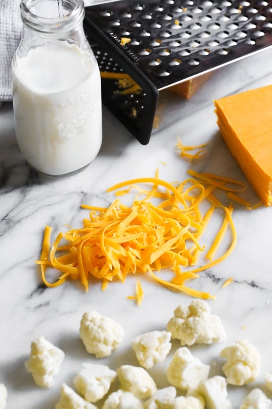 Cheesy cauliflower mac and cheese is perfect when you're craving macaroni and cheese, without the pasta. Each serving is loaded with veggies! Shredded cheddar cheese on a countertop next to cauliflower florets.