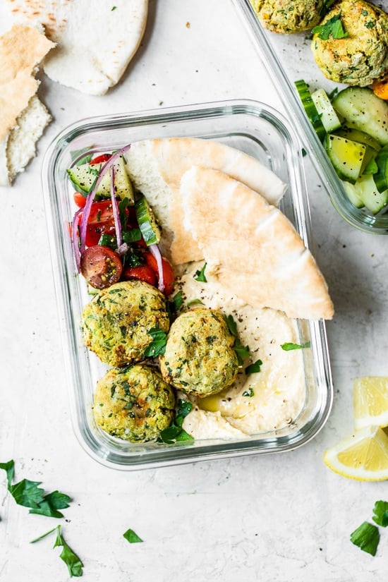 This easy air fryer falafel recipe is made quicker and lighter with canned chickpeas, and no deep frying! Perfect for serving with hummus. Overhead view of homemade falafel in meal prep container with pita bread slices, hummus, and a tomato cucumber salad.