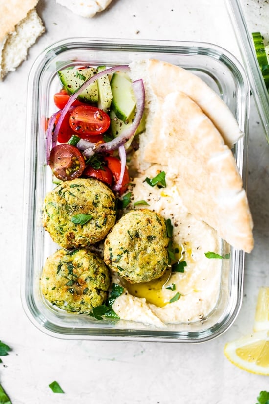 This easy air fryer falafel recipe is made quicker and lighter with canned chickpeas, and no deep frying! Perfect for serving with hummus. Overhead view of homemade falafel in meal prep container with pita bread slices, hummus, and a tomato cucumber salad.