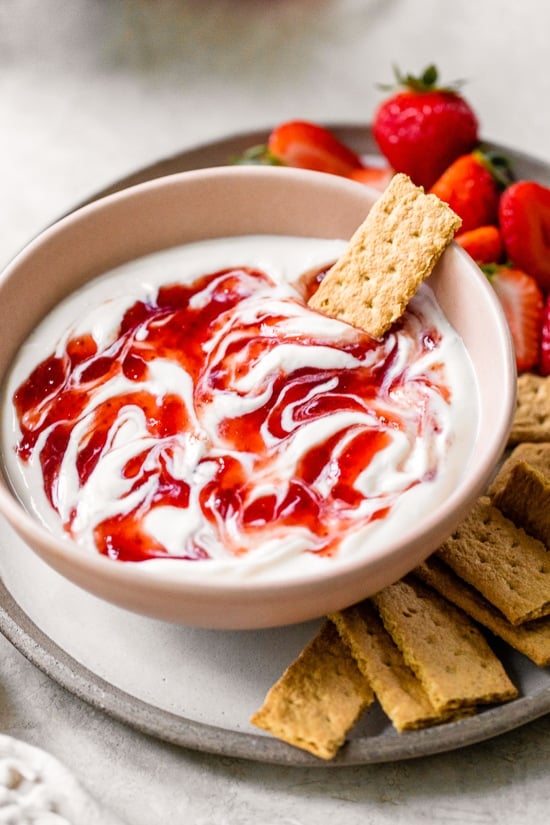 Creamy strawberry cheesecake dip tastes just like cheesecake in dip form! Serve with fresh fruit or graham crackers as a snack or appetizer. A bowl of strawberry cheesecake dip surrounded by mini graham crackers and fresh strawberries.