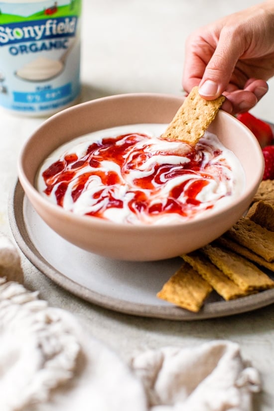 Creamy strawberry cheesecake dip tastes just like cheesecake in dip form! Serve with fresh fruit or graham crackers as a snack or appetizer. A hand dipping a graham cracker into a bowl of cheesecake dip swirled with strawberry preserves.