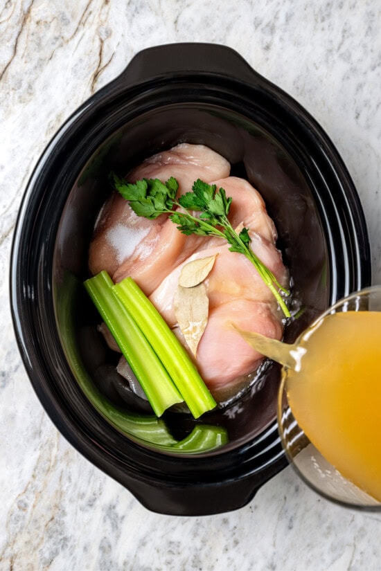 This slow cooker shredded chicken recipe is juicy, tender, and flavorful. It's a hands-off cooking method perfect for meal prep! Pouring broth into slow cooker with chicken breasts, parsley, celery, and bay leaves